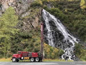 A red drilling truck is parked on a paved area in front of a rocky hillside with a tall, cascading waterfall surrounded by green trees and foliage.