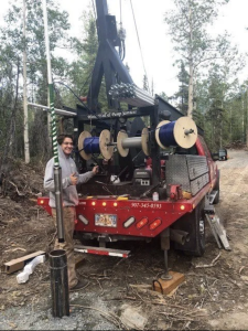 A man stands next to a red utility truck with spools of cable and equipment in a wooded area, smiling and giving a thumbs up. The truck has signs for a pump service company and is parked on a dirt road.