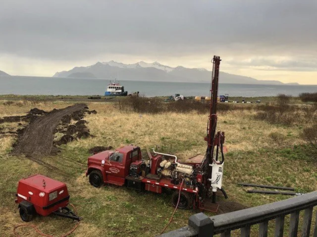 A red drilling truck and generator sit on grassy terrain near a shoreline; a dirt path leads toward water where a boat is docked, with distant mountains visible under a cloudy sky.