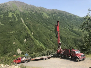 Two red trucks, one with a tall drilling rig, are parked on a gravel area at the base of a lush, green mountain covered in vegetation under a clear sky.