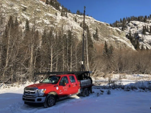 A red pickup truck with company logos is parked on snowy ground in a forested, mountainous area. Attached equipment rises from the truck bed. Snow covers the ground, trees, and rocky hillside under a clear blue sky.