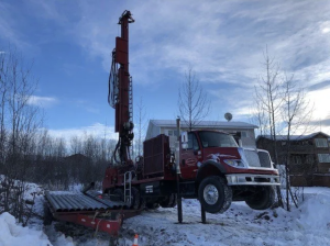 A red drilling truck with its front wheels lifted off the snowy ground uses stabilizing legs while drilling near metal pipes, with houses and bare trees in the background under a blue sky.
