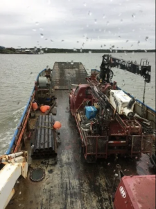 A wet deck of an industrial workboat carrying drilling equipment, pipes, tools, and orange buoys, with water and a distant shoreline visible under a cloudy sky. Raindrops are visible on the camera lens.