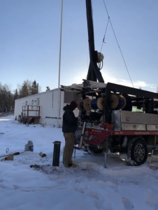 A person stands beside a truck with large spools and equipment in a snowy area, near a white building and trees. The sun is low in the sky, casting long shadows.