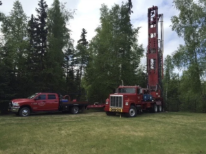 A red pickup truck and a large red drill rig truck are parked on grass near tall green trees under a partly cloudy sky.