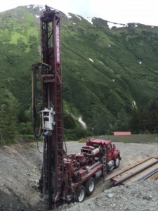 A large red drilling rig truck with SCHRAMM written on its mast is operating on rocky ground, surrounded by green hills and snow-capped mountains in the background.