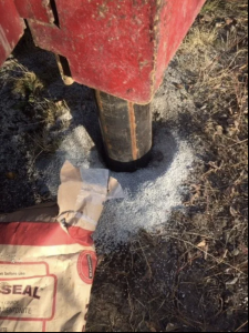 A metal post is set in the ground with gravel around its base. An open bag of bentonite, labeled QUICKSEAL, lies nearby on dry grass and dirt. The red edge of machinery is visible at the top.
