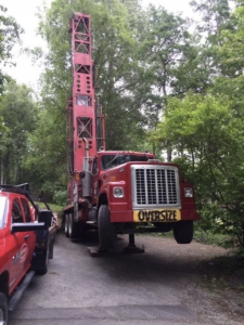 A large red truck labeled OVERSIZE is lifted off the ground by hydraulic stabilizers, with its front wheels elevated. Trees and another red vehicle are visible in the background.