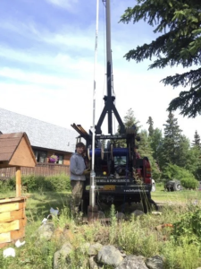 A person operates a well drilling truck in a grassy yard with trees and a house nearby, using equipment to work on a vertical pipe. Tools and supplies are visible around the work area under a clear sky.