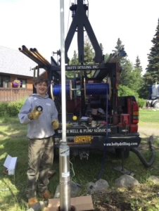 A person wearing work gloves stands next to a metal well pump, smiling and holding a part. Behind them is a drilling truck with equipment and pipes, parked on a grassy yard with trees and a house in the background.
