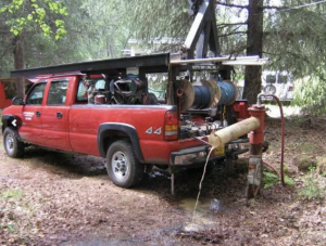 A red pickup truck equipped with water hoses and machinery is parked in a wooded area, connected to a red water hydrant with water flowing out through a pipe into a muddy patch on the ground.
