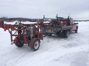 A red pickup truck is parked on snow, towing a trailer with drilling equipment. The background features a snowy field and trees under a gray, overcast sky.
