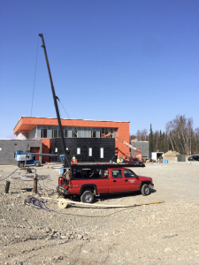A red truck with equipment and a tall crane is parked on a gravel lot in front of a building under construction, with workers, ladders, and scaffolding visible; trees and clear sky in the background.