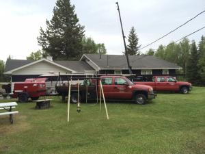 Two red utility trucks are parked on the grass in front of a house with a gray roof. Equipment, a swing set, and a picnic table are also in the yard, surrounded by trees under a cloudy sky.