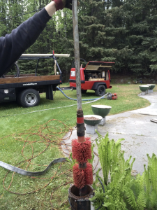 A person holds a metal pipe with red bristle brushes attached to it above a well opening outdoors; work trucks, hoses, and tools are visible on the grass in the background.