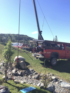 A red pickup truck with its tailgate open is parked on grass near rocks. A crane extends upward behind the truck, and ropes are attached to a nearby tree. Hills and trees are visible in the background under a clear sky.