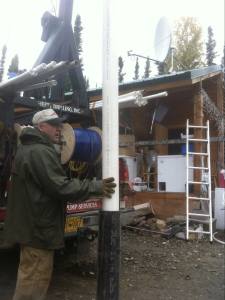 An older man in a green jacket and cap holds a large white PVC pipe upright near a truck and a rustic building with tools, a ladder, and satellite dish visible in the background.