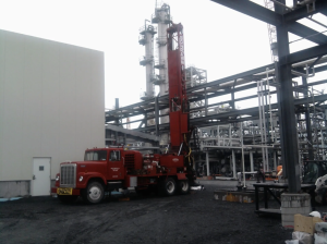 A red drilling truck is parked at an industrial construction site with metal structures and a tall processing tower in the background under an overcast sky.