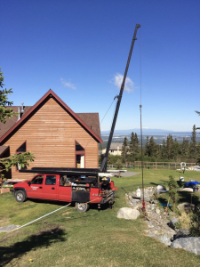 A red utility truck with equipment is parked next to a wooden house, supporting a tall crane lifting an object in a grassy yard with rocks. Trees, a fence, and mountains are visible in the background under a clear blue sky.