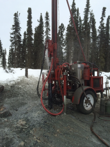 A red drilling rig operates on snowy, forested ground, spraying water as it drills into the earth; tall pine trees stand in the background under an overcast sky.
