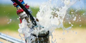 Close-up of water spraying forcefully from a metal pipe joint outdoors, with droplets captured mid-air and blurred greenery in the background.