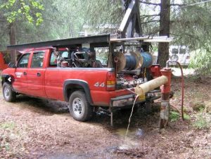 A red pickup truck fitted with reels and equipment is parked in a wooded area, connected by a large hose to a water well hydrant, indicating it is being used for water extraction or well maintenance.