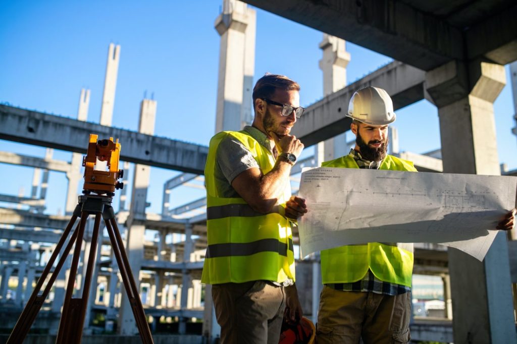 Two construction workers in safety vests and helmets examine blueprints at a building site with concrete columns and beams under a clear blue sky. A surveying instrument is visible in the foreground.