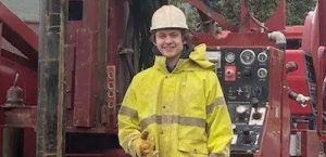 A person wearing a yellow rain jacket, white hard hat, and gloves stands in front of industrial machinery with control panels, smiling at the camera.