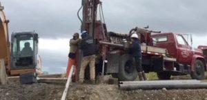 Several workers in safety gear operate drilling equipment next to a red truck at a construction site under a cloudy sky. A worker sits in an excavator on the left, while pipes lie on the ground in the foreground.