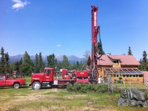 A red drilling rig truck is parked beside a log cabin with a greenhouse. Mountains and pine trees are in the background under a clear blue sky on a sunny day.