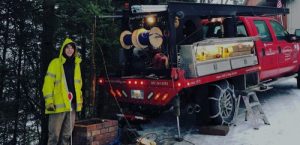 A utility worker in a yellow jacket stands next to a red service truck equipped with tools and cable reels. Snow covers the ground, and the truck is parked near trees and a brick structure.