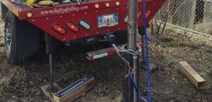 A red drilling truck with extended stabilizers is parked on dry ground. Equipment and tools are mounted at the back, with cables and hoses connected to a metal pipe in the ground. Wooden blocks and leaves are scattered nearby.