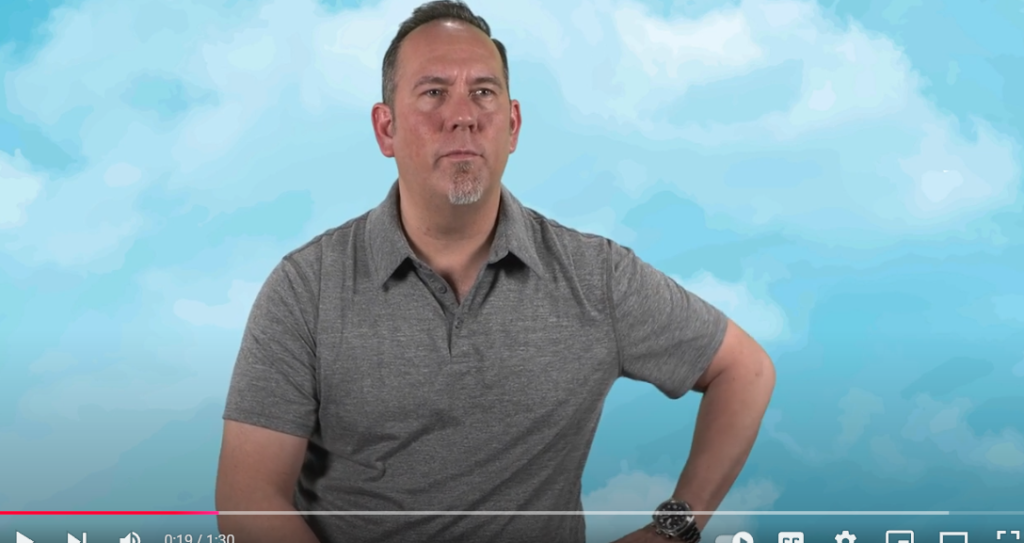 A man wearing a gray polo shirt sits with one hand on his hip against a blue sky and clouds background. He looks ahead with a neutral expression. Video player controls are visible at the bottom of the image.