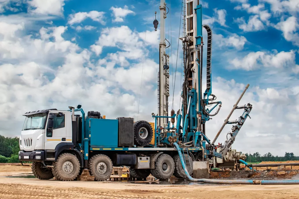 A large blue and white well drilling truck is parked on a construction site, equipped with drilling machinery and hoses, under a partly cloudy sky with open land in the background. - Water Well Drilling | Hefty Drilling Professional well drilling contractor using modern equipment at Alaska drilling site